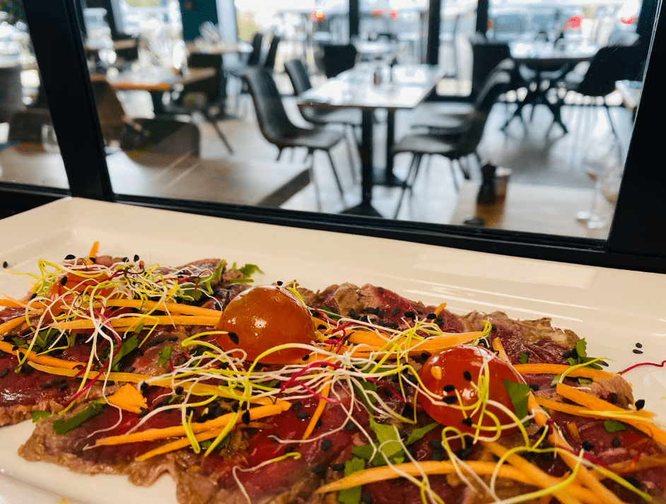 Carpaccio de bœuf ou thon frais sur assiette blanche, garni de pousses, carottes râpées et tomates cerises, avec restaurant en arrière-plan flou.